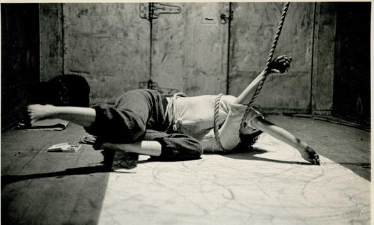 An image of Carolee lying on the floor with her arms tied up in some rope 