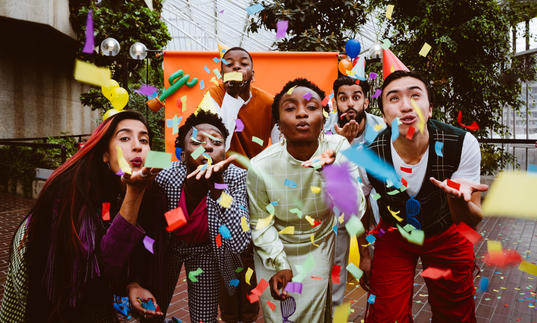 The PappyShow performers blow multi coloured confetti through the air towards the camera. 