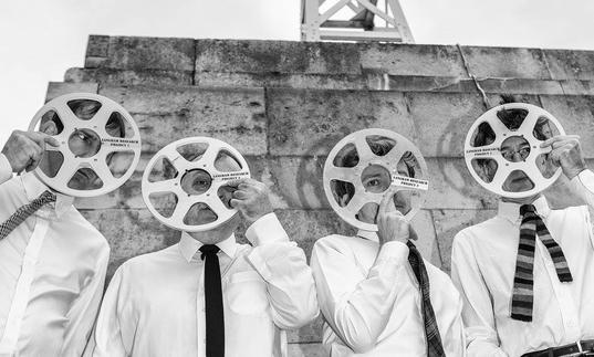 A black and white image of four men dressed in shirts and ties, holding tape reels over their faces