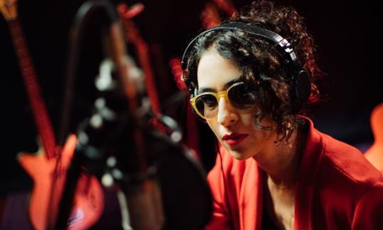 a close up photo of Marisa Monte wearing sunglasses and a red suit against a dark background