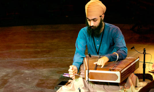 Eeshar Singh playing the santoor - a flat stringed instrument. He is wearing a beige turban and blue shirt.