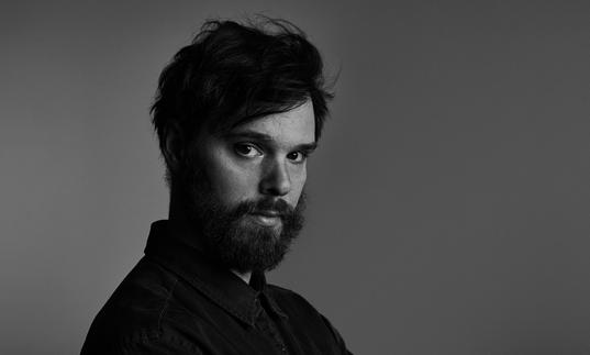 A black and white portrait of Dave Longstreth, looking sideways at the camera. He is wearing a dark shirt and has a beard.