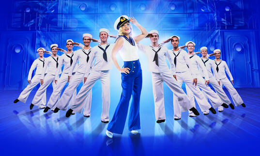 Kerry Ellis dressed in a nautical themed outfit and hat as Reno Sweeney. She stands surrounded by sailors, all saluting.