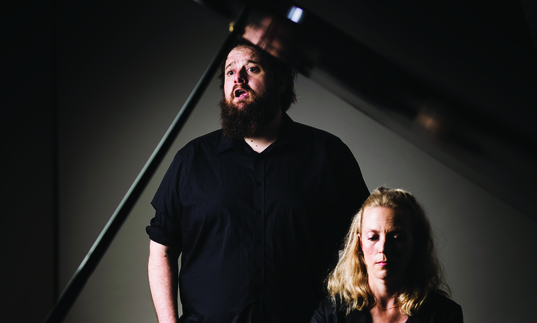 Allan Clayton singing as he stands behind a piano accompanist