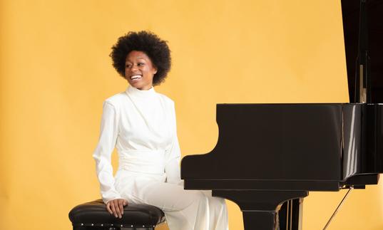 Isata Kanneh-Mason sitting at a grand piano smiling against a pale orange backdrop