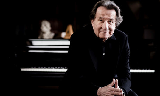 Rudolf Buchbinder sits with his hands touching, with a piano keyboard blurred out behind him.