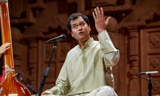 Uday Bhawalkar singing with his left hand raised. He is sitting cross-legged wearing a pale green tunic.