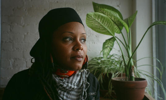 Matana Roberts looking out a window with a plant in front of her. She is wearing a black cap backwards and a white and black dogtooth scarf.