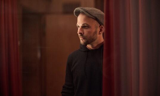 Nils Frahm standing between two red curtains, wearing a flat cap with a black hoodie