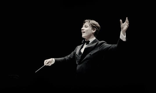 Klaus Makela is conducting in front of a black background, his right hand holding the baton and his left hand above his shoulder
