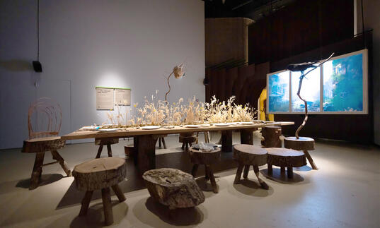 Photograph of artwork Refuge for Resurgence by Superflux showing a wooden table where creatures can sit.