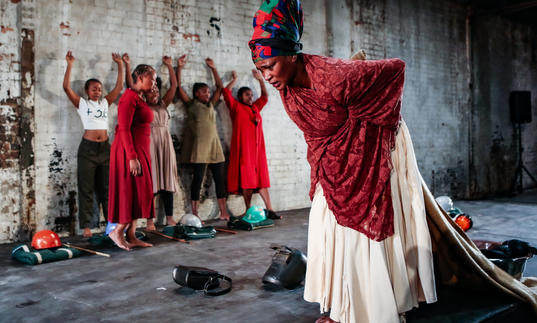 A performer in a cream dress and red shawl moves across the stage, behind her a group of 5 performers stand with their arms raised against the wall.