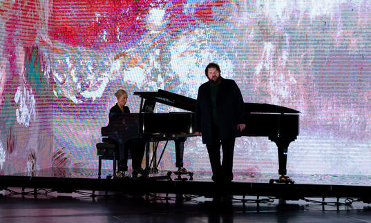 Allan Clayton stands in front of a piano at which Kate Golla sits, with a pink landscape by Fred Williams OBE projected on a screen behind them