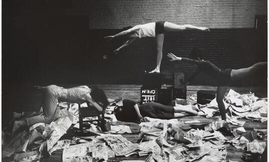 Image shows 4 dancers posing horizontally amongst newspapers 