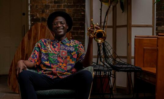 Mark Kavuma sitting laughing in a chair, laughing, holding a trumpet in a brightly patterened shirt and brown hat