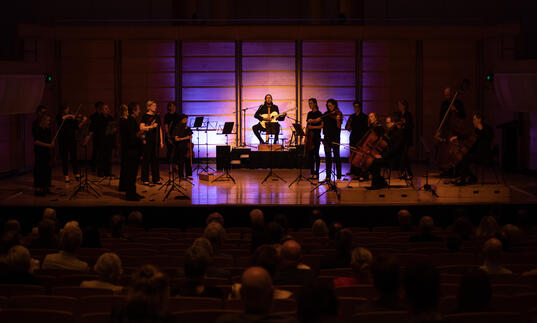 William Barton sits in the middle of the stage on a pedestal playing guitar and didgeridoo. Members of the ACO stand either side of him, in near-darkness