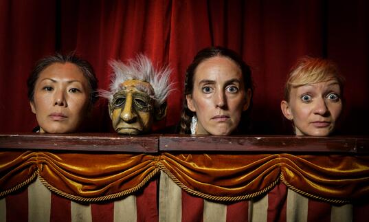 Three performers and one puppet stand in a puppet booth and stare directly at the camera.