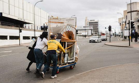 image of three people pushing a trolley with furniture up a hill with buildings and cars in the background