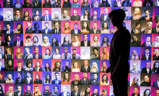 Digital installation of portraits of hundreds of people on a pink or purple ton background with the silhouette of a person standing in front of the installation on the right. 