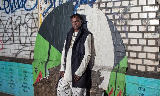 Dynamic image of Baaba Maal, standing against a graffiti covered brick wall