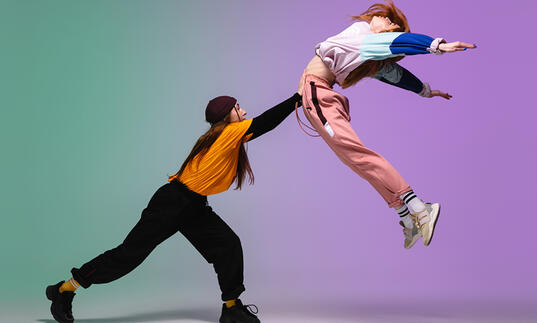 Two girls dancing, one with arm outstretched, pushing the other into the air, against a pastel coloured background