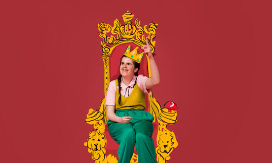 A photo of Rachel sitting on an illustrated golden throne wearing an illustrated golden crown. She is wearing green trousers and a pink top with a yellow top on top of it.  She is smiling with her finger pointed towards the sky.