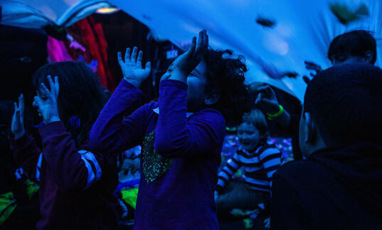 A group of children gather underneath a large blue sheet. They laugh, smile and lift their hairs upwards towards it.