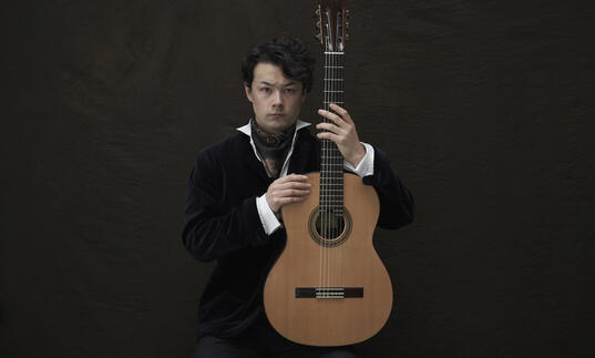 Sean Shibe sitting in front of a dark grey background, holding his guitar upright