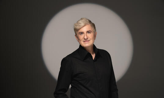 Jean Yves Thibaudet standing in front of a grey background with a spotlight forming a light circle behind him