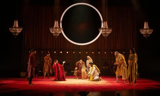 An ensemble of performers gather on stage in a decadent room, a large circle light hangs above them.