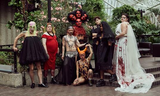 Transpose Burn artists standing in the Barbican Conservatory surrounded by plants. From the left we have Kübra Uzun (they, them),  Kell w Farshéa (they, them), Mustafa Kınalı (aka Mustkika) (any pronouns), Akış Ka (they, them), Dani Dinger (any pronouns),CN Lester (they, them), Ifeoluwa (they, them) and i-Gemini (he, him)