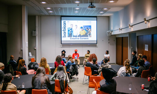 Group of people sitting in a room facing a screen and 4 people people sitting on a panel 