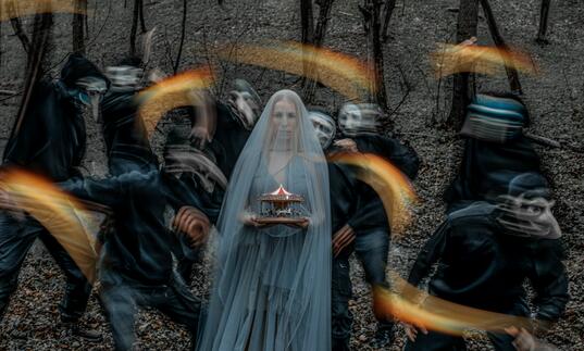A lady in long kthin light blue veil is staring at the camera. She is holding a small model of a red and white carousel in her hands. Behind her seven masked figure and blurred as they move, they are holding flares.