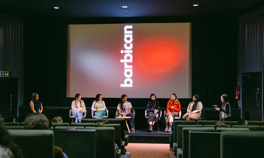 image of a panel seated in front of a cinema screen which has the Barbican logo displayed