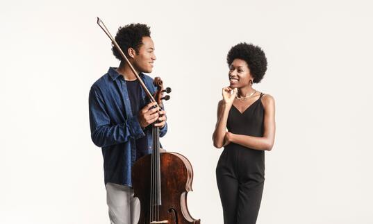 Sheku and Isata Kanneh-Mason smiling at each other. Sheku is holding his cello and bow and they are standing in front of a white background.