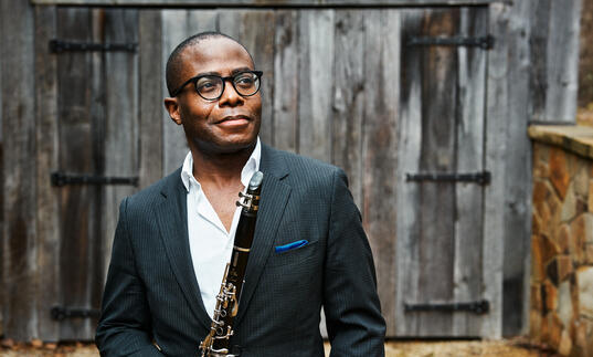 Anthony McGill standing in front of a wooden barn holding his clarinet