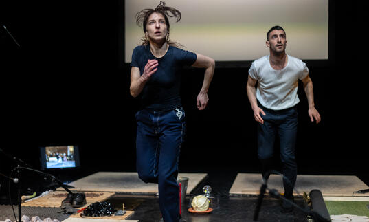 Two dancers run on treadmills... they look out at the audience, watching something, concerned.