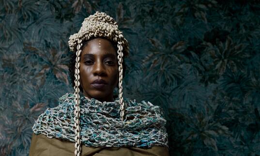 Performer Julene Robinson stands against a blue floral background. She wears cream and blue chains around her head and shoulders.