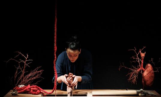 A person wearing blue overalls sculpts a small body out of clay on a table. There is a heart to their left-hand side and a red rope that mimics an intestine.