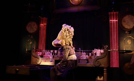 A person with a large nose as a head sits at a table that is filled with perfume bottles.