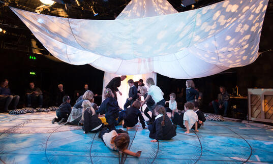 A group of families enjoying a performance of Told by an Idiot's Get Happy. Children gather under a big, white parachute. 