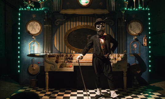 A person dressed as an insect with antennae on their head, wearing a suit, a top-hat and carrying a cane, stands in front of an organ.