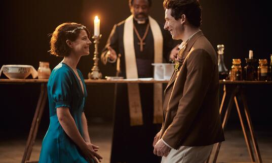 Romeo and Juliet marry, smiling hugely at each other in front of Friar Lawrence, in a candlelit hall. 
