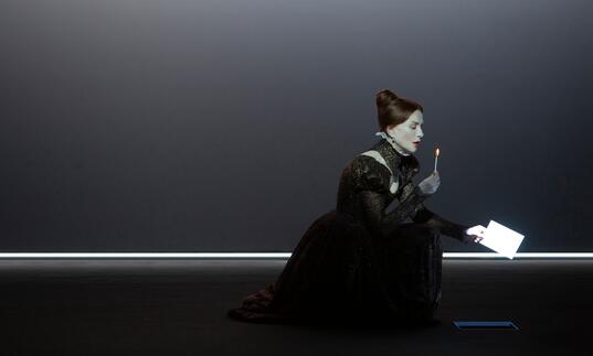 A person dressed as Mary Stuart kneels on the floor in an old-fashioned gown with a lighted match in their hand and a letter in the other.