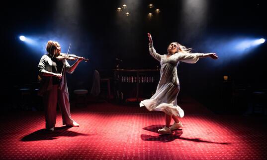 A person in a white dress dances next to a person playing the violin. They are in a dark space, dressed up like a pub with wooden tables and chairs and a red carpet. 
