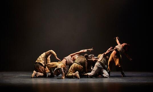 Members of the Boy Blue dance theatre company perform against a black background, one person is standing arching backwards while others kneel and crawl on the floor.