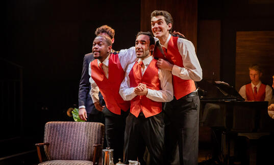 Three singers in red waistcoats, with grand piano in background