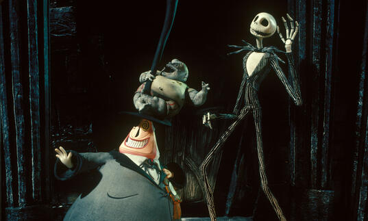 A skeleton man and a short man in a suit dance together on a dark stage.