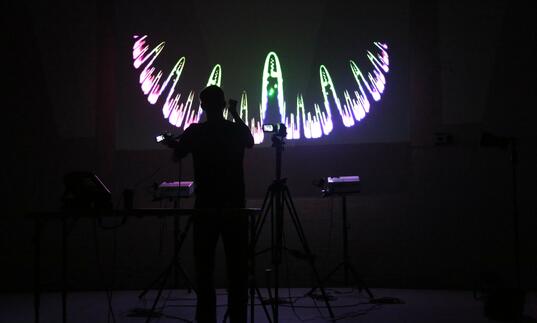 A person stands in a dark room surrounded by technical equipment. A swirling purple and green line is projected before them on a screen.