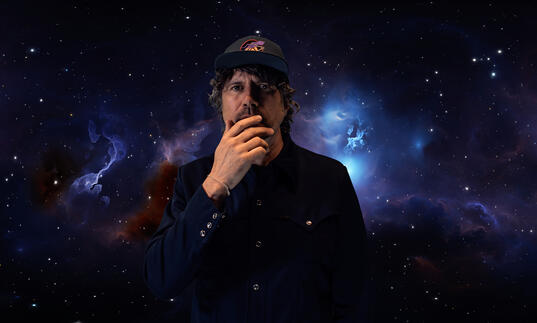 Gruff Rhys stands, hand over mouth in front of galaxy backgrounds
